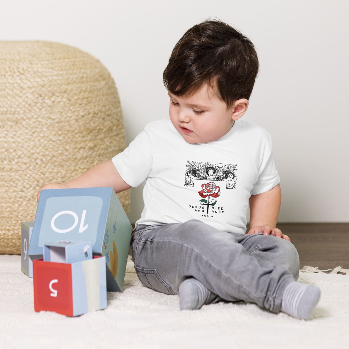 Child sitting on a carpeted floor with a toy and a white Christian t-shirt with graphics and text.
