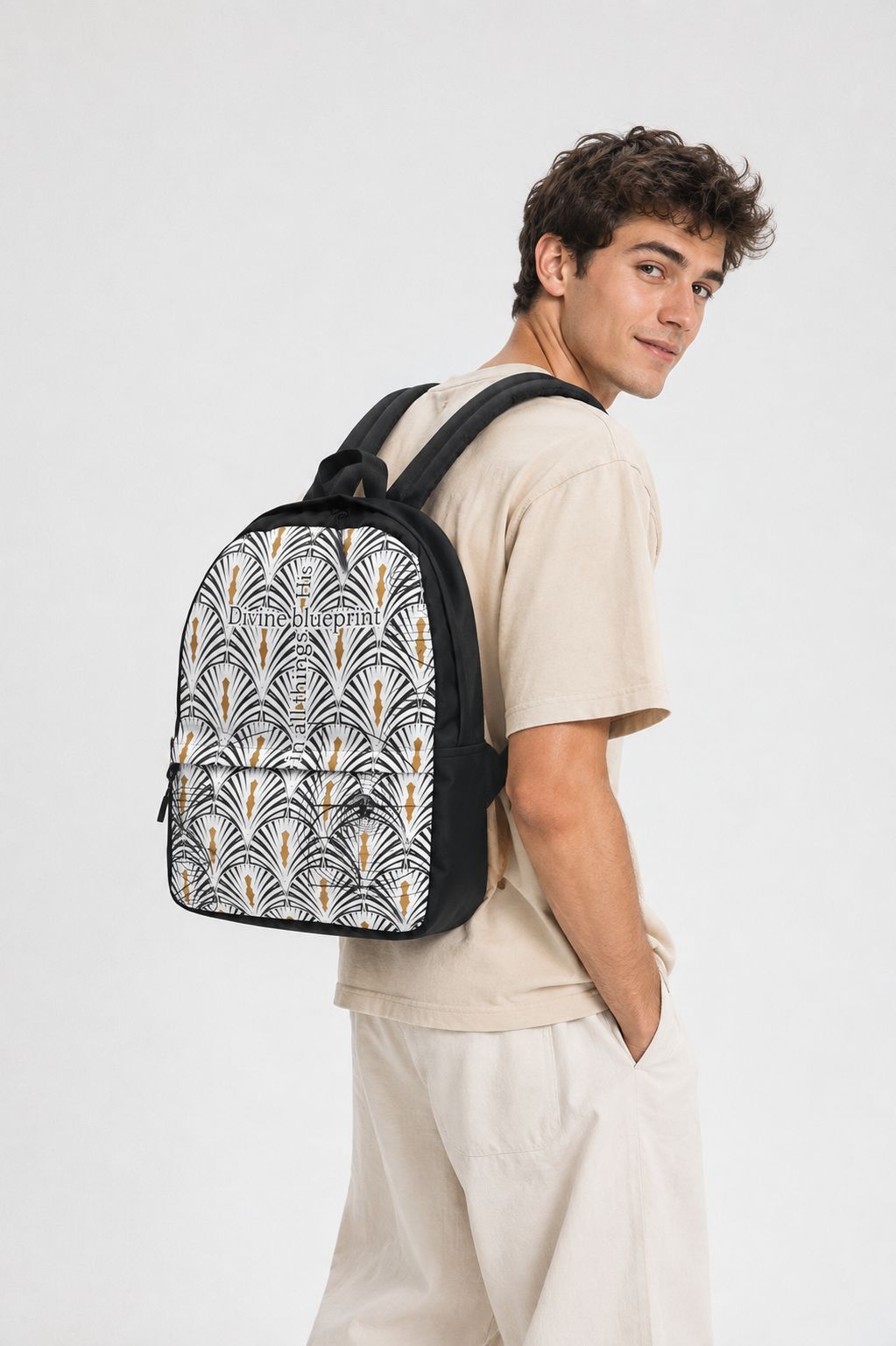 A man wearing a beige t-shirt and light beige pants is shown from the back and side, carrying a black backpack with a white, black, and gold palm leaf pattern against a light gray studio background.