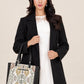 A woman wearing a long black coat over a white lace-trimmed dress, holding a patterned tote bag with black handles against a plain beige studio background.