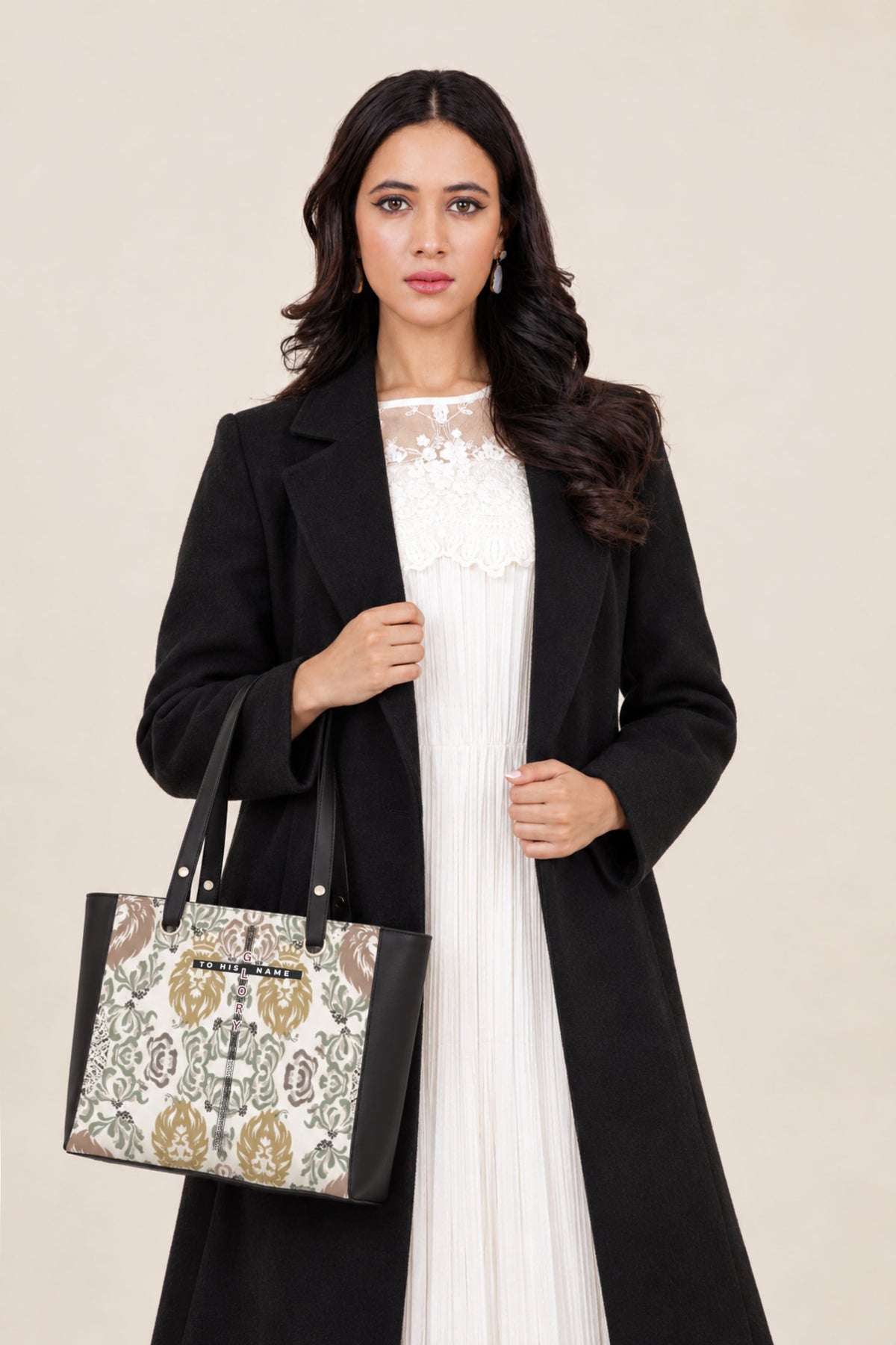 A woman wearing a long black coat over a white lace-trimmed dress, holding a patterned tote bag with black handles against a plain beige studio background.