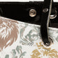 Close-up of a tote bag featuring a white base with a pattern of stylized lion heads in beige, gray, and mustard yellow, accented by a glossy black trim and strap with silver-tone metal hardware.