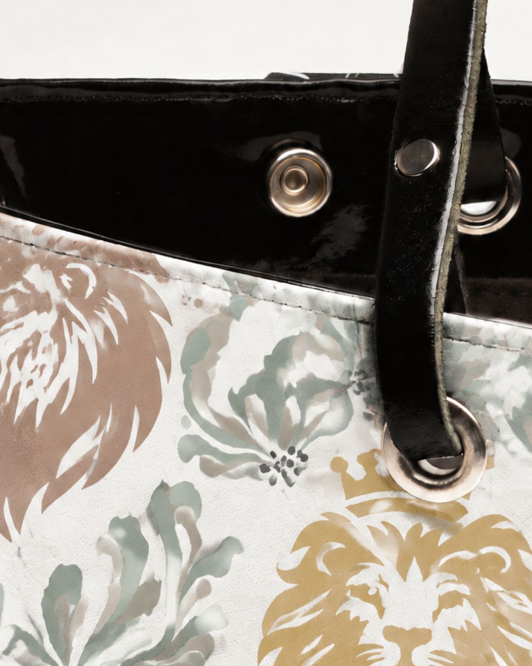 Close-up of a tote bag featuring a white base with a pattern of stylized lion heads in beige, gray, and mustard yellow, accented by a glossy black trim and strap with silver-tone metal hardware.