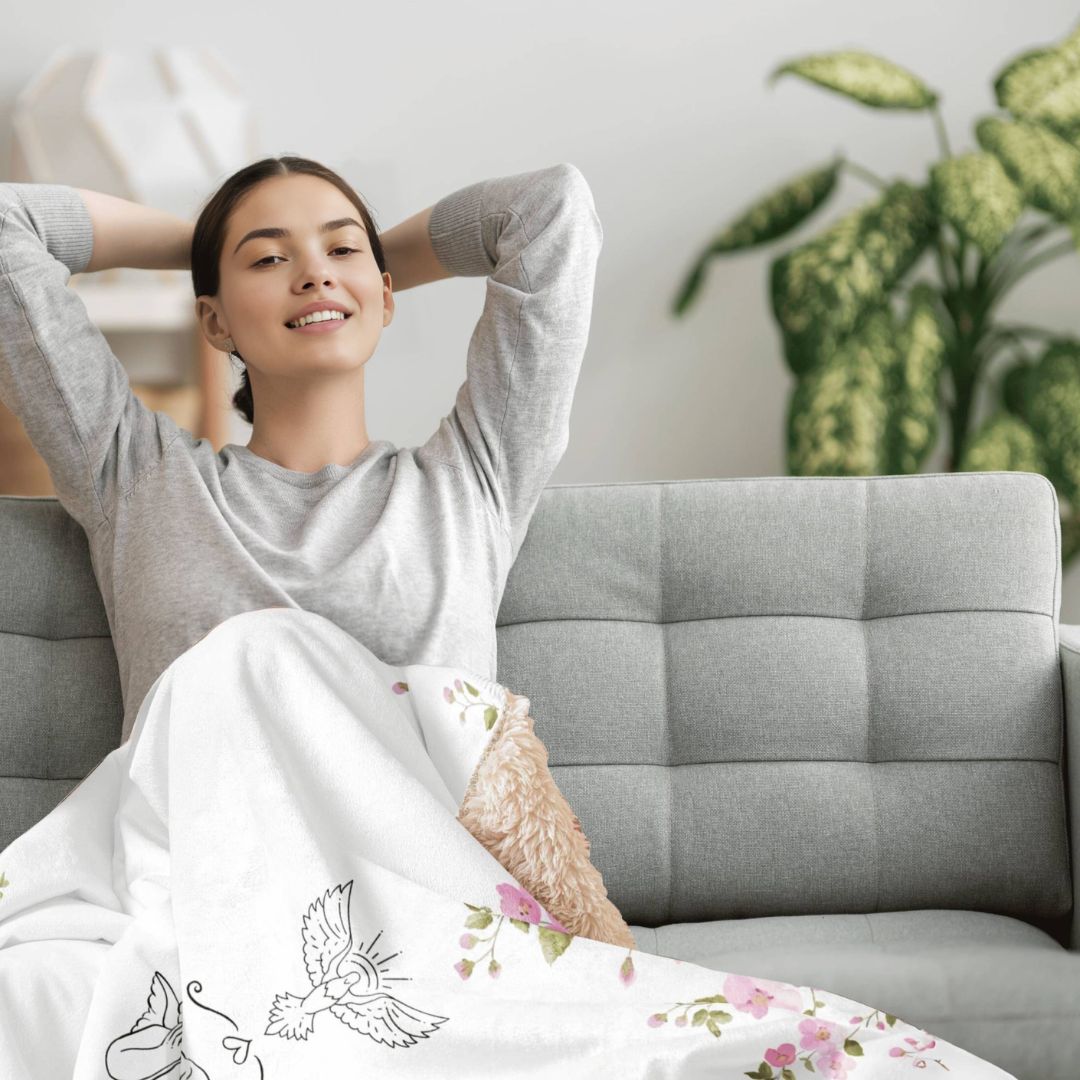 A person relaxing on a couch, covered with a sherpa blanket featuring a floral design with ornaments in the center.