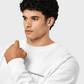 A man with curly black hair wearing a white crewneck sweatshirt with black text on the left chest, photographed against a light gray studio background.