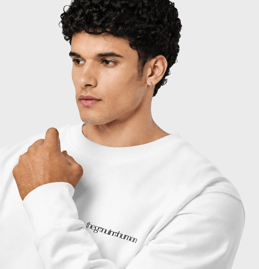 A man with curly black hair wearing a white crewneck sweatshirt with black text on the left chest, photographed against a light gray studio background.