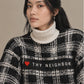 A woman wearing a black and white plaid knit sweater with a white ribbed turtleneck underneath. The sweater features a black band across the chest with the text 'THY NEIGHBOR' and a small red heart icon.