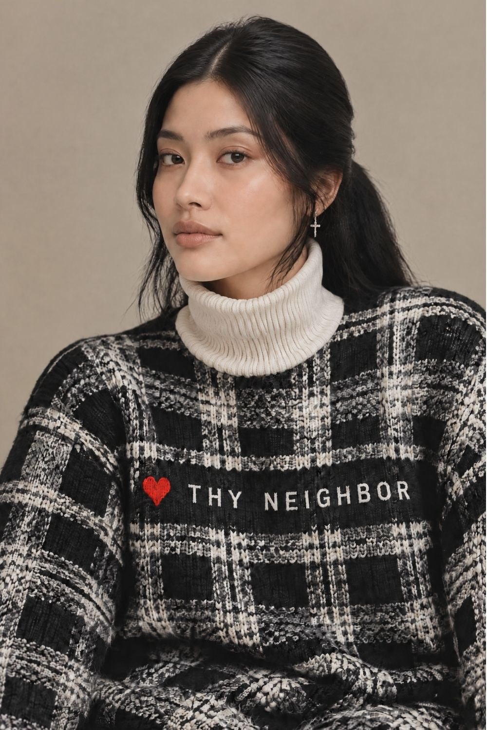 A woman wearing a black and white plaid knit sweater with a white ribbed turtleneck underneath. The sweater features a black band across the chest with the text 'THY NEIGHBOR' and a small red heart icon.