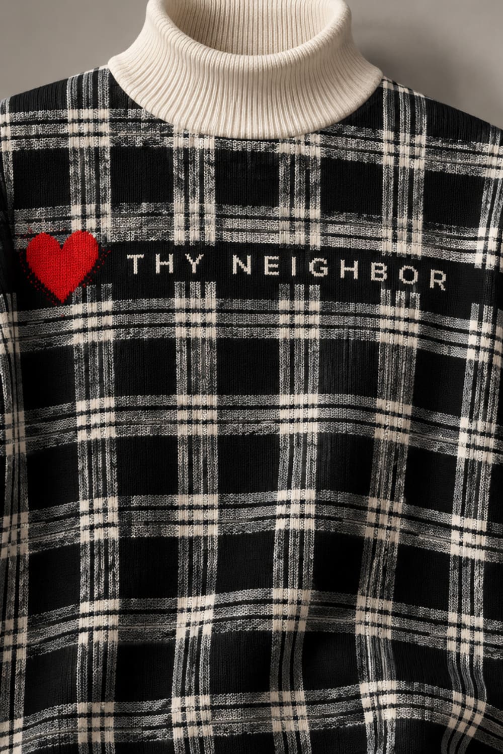 A close-up of a black and off-white plaid turtleneck sweater featuring a red heart and the text 'THY NEIGHBOR' across the chest.