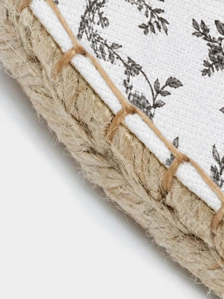 Close-up of a woven textile product showing a white fabric base with a black floral toile pattern and a thick braided natural jute rope trim bound by tan thread.