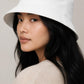 A woman wearing a white bucket hat with a black embroidered cross and 'THE WAY TRUTH LIFE' text on the front, paired with a beige knit sweater against a neutral beige background.