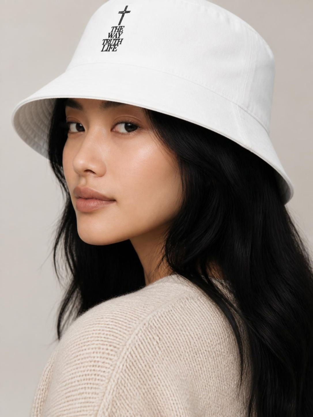 A woman wearing a white bucket hat with a black embroidered cross and 'THE WAY TRUTH LIFE' text on the front, paired with a beige knit sweater against a neutral beige background.