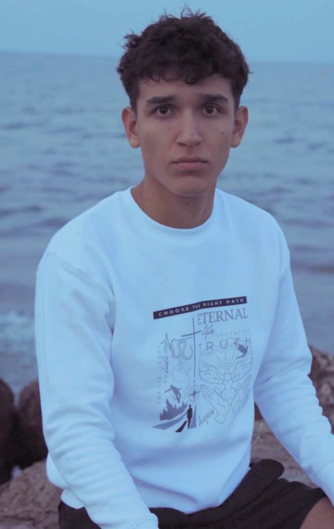 Young man sitting on a stone with the ocean behind him and wearing a white Christian faith sweatshirt water 